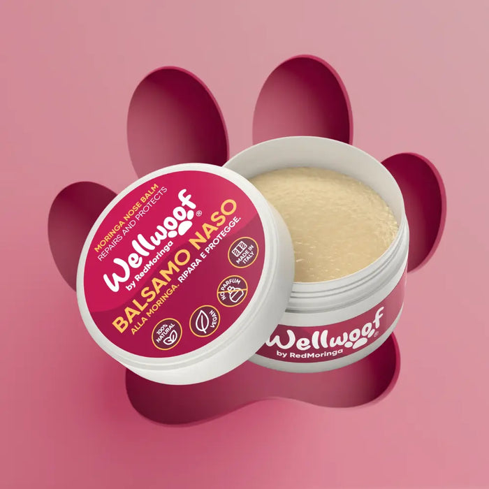 Primo piano del barattolo di Balsamo Naso Wellwoof by RedMoringa su uno sfondo rosa con l'ombra di un'impronta di zampa. Il packaging evidenzia che il prodotto è 100% naturale, vegano e senza profumo.